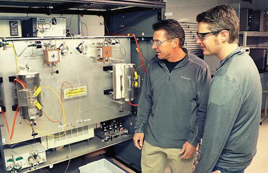 Troy Semelsberger (left) and Andrew Sutton stand alongside the continuous flow reactor that their team developed. The high-throughput environment could improve biofuels production by enabling more efficient chemical conversion of biomass into hydrocarbon fuels.